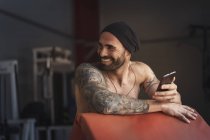 Atleta tatuata senza maglietta ridente con smartphone in palestra — Foto stock