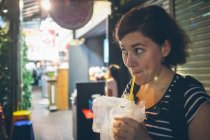 Смішна жінка-мандрівник смокче свіжий напій з пляшки і озирається геть, стоячи біля кафе на міській вулиці — стокове фото