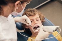 Femme en masque et uniforme de médecin faisant le balayage des dents du petit garçon tout en travaillant dans la clinique dentaire — Photo de stock