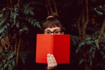 Молода елегантна жінка в окулярах дивиться на камеру, прикриваючи обличчя книгою в міському саду — стокове фото