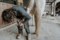 Неузнаваемый взрослый мужчина с помощью ножа удаляет грязь с копыт лошади на ранчо — стоковое фото
