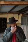 Adult male in hat smoking cigarette while standing near stalls inside stable on ranch — Stock Photo