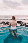 Vista trasera de la mujer en traje de baño descansando en el agua de la piscina cerca de rocas y cielo nublado en la costa del mar - foto de stock
