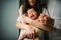 Hombre y mujer anónimos abrazando y reconfortando llorando bebé recién nacido en la habitación acogedora en casa - foto de stock