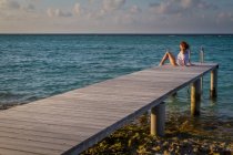 Вид сбоку на красивую молодую женщину, смотрящую в сторону, сидя на деревянном пирсе возле волнистой морской воды на фоне облачного вечернего неба на Мальдивах — стоковое фото