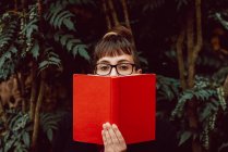Jeune femme élégante avec des lunettes regardant la caméra tout en couvrant le visage avec un livre dans le jardin de la ville — Photo de stock