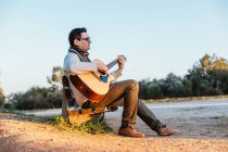 Casual uomo in occhiali suonare la chitarra su panchina in campagna — Foto stock