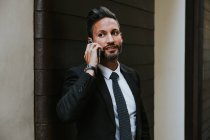 Adult handsome elegant businessman in formal suit looking away and talking on mobile phone near wall — Stock Photo