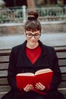 Giovane donna elegante sorridente in occhiali libro di lettura e seduta sulla panchina nel parco cittadino — Foto stock
