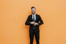 Adulto bonito homem de negócios elegante em terno formal ajustando jaqueta e olhando para a câmera perto da parede laranja — Fotografia de Stock