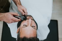 Primer plano del peluquero con peine y recortadora cortando la barba del macho sentado en la peluquería sobre fondo borroso - foto de stock