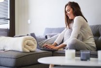 Веселая молодая женщина использует ноутбук и отдыхает на диване дома — стоковое фото