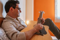 Чоловік в пуловері грає на гітарі на дивані вдома — стокове фото