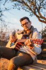 Случайный мужчина в очках, играющий на гитаре на скамейке в сельской местности — стоковое фото