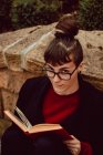 Junge elegante Frau mit Brille lehnt mit Buch an Steinmauer und blickt in die Kamera — Stockfoto