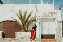 Nachdenkliche junge Frau lehnt an schäbiger Hauswand in der Nähe exotischer Palmen an sonnigem Tag — Stockfoto