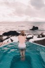 Вид ззаду жінки в купальнику, що відпочиває у воді басейну біля скель і хмарного неба на морському узбережжі — стокове фото