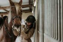 Vue latérale de la jeune femelle en casque mettant la selle sur le cheval merveilleux près de l'écurie sur ranch — Photo de stock
