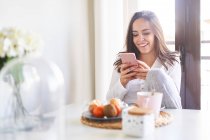 Attrayant jeune femme heureuse en utilisant le téléphone portable et prendre le petit déjeuner à table près de la fenêtre à la maison — Photo de stock