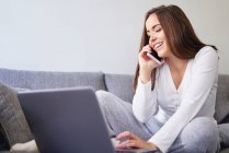 Улыбающаяся молодая счастливая женщина с помощью ноутбука и говорить по мобильному телефону на диване дома — стоковое фото