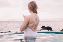 Young woman meditating in water of pool near rocks and cloudy sky — Stock Photo