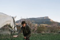 Старик в шляпе смотрит в сторону и сидит на красивой лошади против безоблачного голубого неба на лугу — стоковое фото
