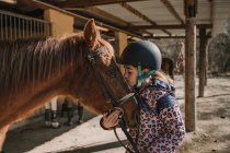 Cute little girl in helmet kissing a white horse while standing near stalls in stable during horseback riding lesson on ranch — Stock Photo