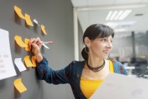 Mujer de negocios sosteniendo documento y escribiendo en notas adhesivas mientras está de pie cerca de la pared gris en la oficina - foto de stock