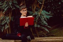 Jovem mulher elegante em óculos ler livro e sentado no banco no jardim da cidade — Fotografia de Stock