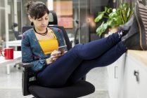 Pesebre femenino usando teléfono inteligente mientras mantiene las piernas en el escritorio en la oficina - foto de stock
