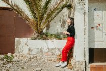 Vue latérale d'une charmante jeune femme aux yeux fermés appuyée sur un mur minable d'une vieille maison près d'un palmier exotique par une journée ensoleillée — Photo de stock