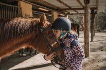 Menina bonito no capacete beijando um cavalo branco enquanto está perto de barracas no estábulo durante a aula de equitação no rancho — Fotografia de Stock