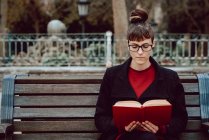 Junge attraktive elegante Frau mit Brille liest Buch und sitzt auf Bank im Stadtgarten — Stockfoto