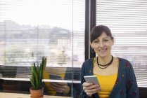 Портрет жінки-менеджера, який тримає смартфон біля вікна під час роботи в сучасному офісі — стокове фото