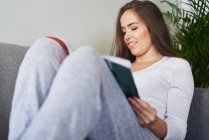 Молода щаслива жінка читає і відпочиває на дивані вдома — стокове фото
