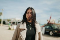 Jovem mulher pensativa em roupa da moda com jaqueta em pé no fundo borrado de estacionamento na praia — Fotografia de Stock