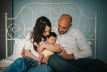 Mère et père heureux embrassant bébé nouveau-né adorable tout en étant assis sur un lit confortable ensemble — Photo de stock