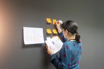 Geschäftsfrau hält Dokument in der Hand und schreibt auf klebrige Zettel, während sie in der Nähe einer grauen Wand im Büro steht — Stockfoto