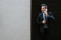 Adulto bonito homem de negócios elegante em terno formal olhando para longe e falando no telefone móvel perto da parede — Fotografia de Stock