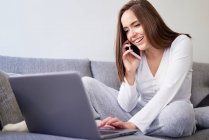 Smiling young happy woman using laptop and talking on mobile phone on sofa at home — Stock Photo