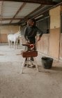 Farrier adulto usando martillo y pinzas para forjar herradura caliente en yunque portátil cerca de establo en rancho - foto de stock