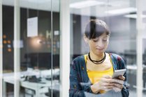 Manager femminile sorridente utilizzando smartphone mentre in piedi vicino alla parete di vetro in ufficio moderno — Foto stock