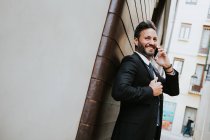 Дорослі красиві елегантний бізнесмен в офіційному костюмі, дивлячись на нього і розмовляючи по мобільному телефону біля стіни — стокове фото