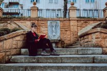 Giovane donna elegante in occhiali da vista lettura libro e seduto sulle scale di fronte alla costruzione in città — Foto stock