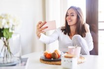 Молода щаслива жінка бере селфі з мобільним телефоном, снідаючи біля вікна вдома — стокове фото