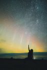 Silhueta de mulher com a mão levantada fazendo ioga em terra perto de incríveis luzes do norte no céu com muitas estrelas à noite — Fotografia de Stock