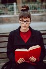 Молода елегантна жінка в окулярах читає книгу і сидить на лавці в міському парку — стокове фото