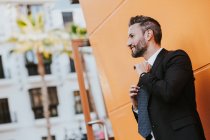 Adulto bonito homem de negócios elegante em terno formal ajustando gravata e olhando para longe perto da parede laranja — Fotografia de Stock
