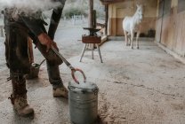 Contadino adulto con martello e pinze per forgiare ferro di cavallo caldo su incudine portatile vicino alla stalla nel ranch — Foto stock