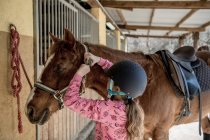 Carino bambina in casco mettere spazzolatura un bianco cavallo mentre in piedi vicino bancarelle in stalla durante equitazione lezione di equitazione su ranch — Foto stock